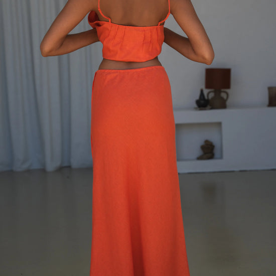 Person wearing an orange outfit standing in a room with a white curtain and lamp in the background.