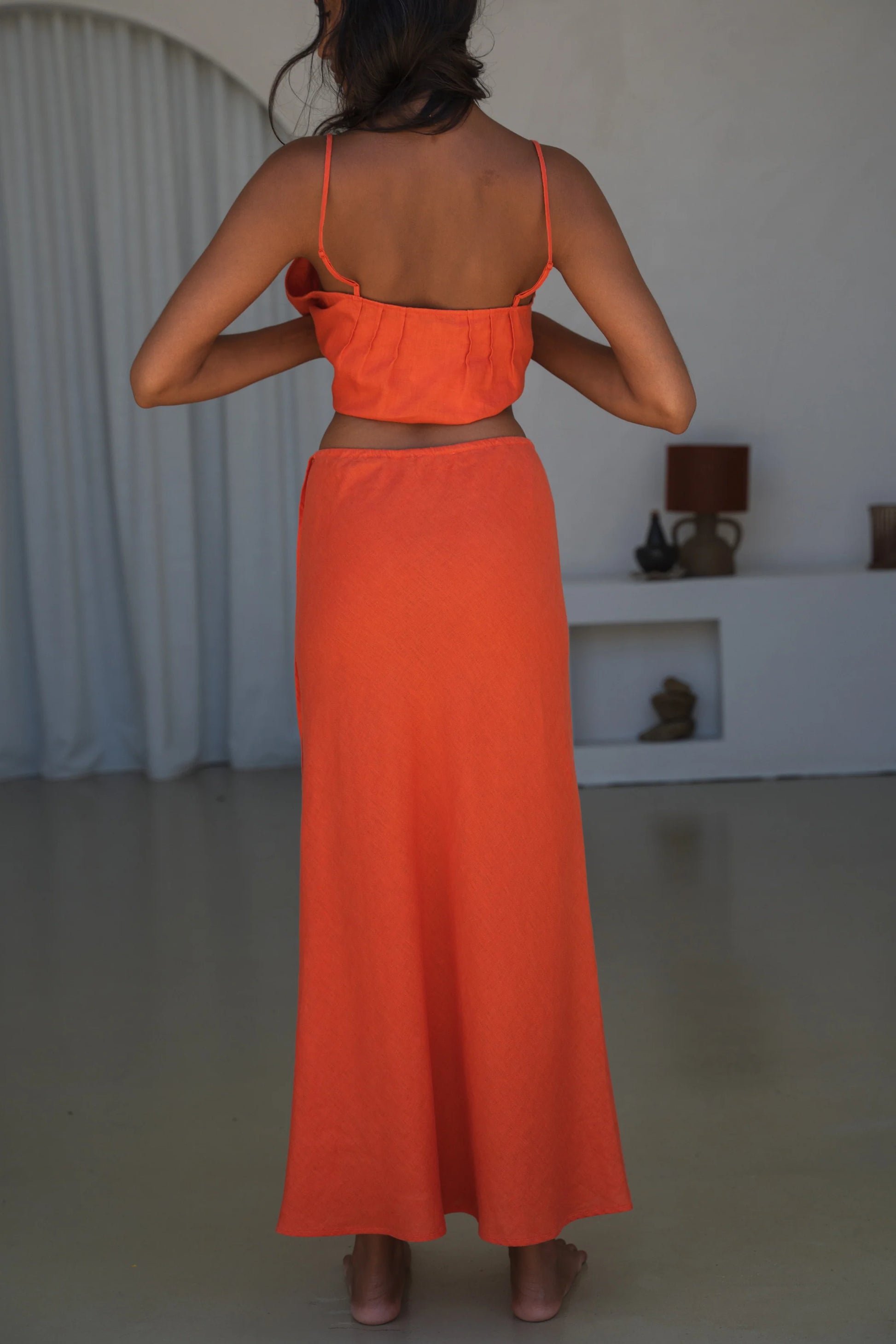 Person wearing an orange outfit standing in a room with a white curtain and lamp in the background.