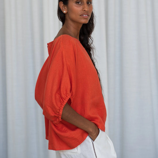 Woman wearing an orange top and white pants against a light curtain background from Lilly Pilly