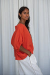 Woman wearing an orange top and white pants against a light curtain background from Lilly Pilly