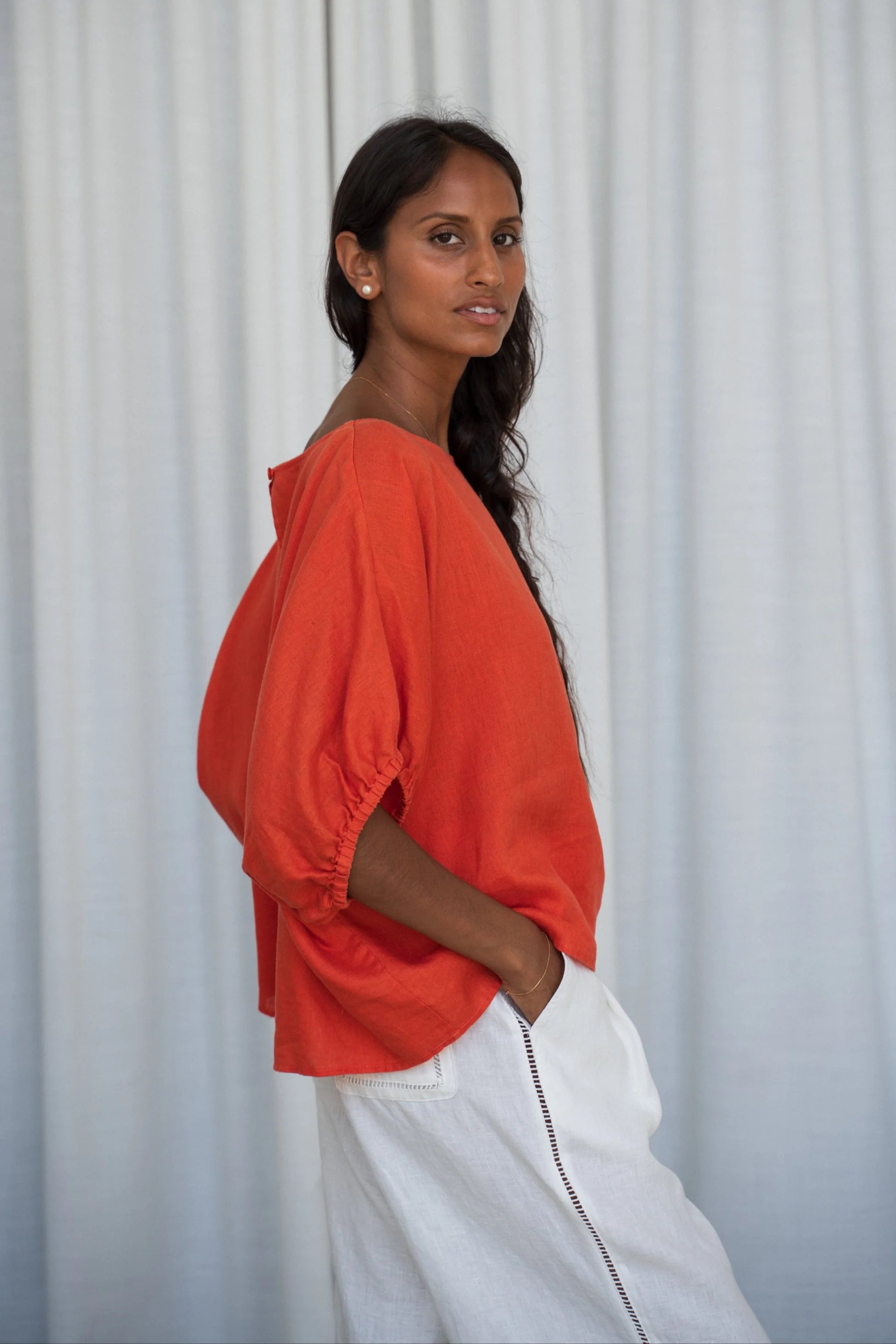 Woman wearing an orange top and white pants against a light curtain background from Lilly Pilly