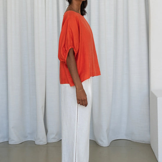 Woman wearing an orange top and white pants standing against a white curtain from Lilly Pilly