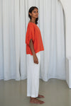 Woman wearing an orange top and white pants standing against a white curtain from Lilly Pilly