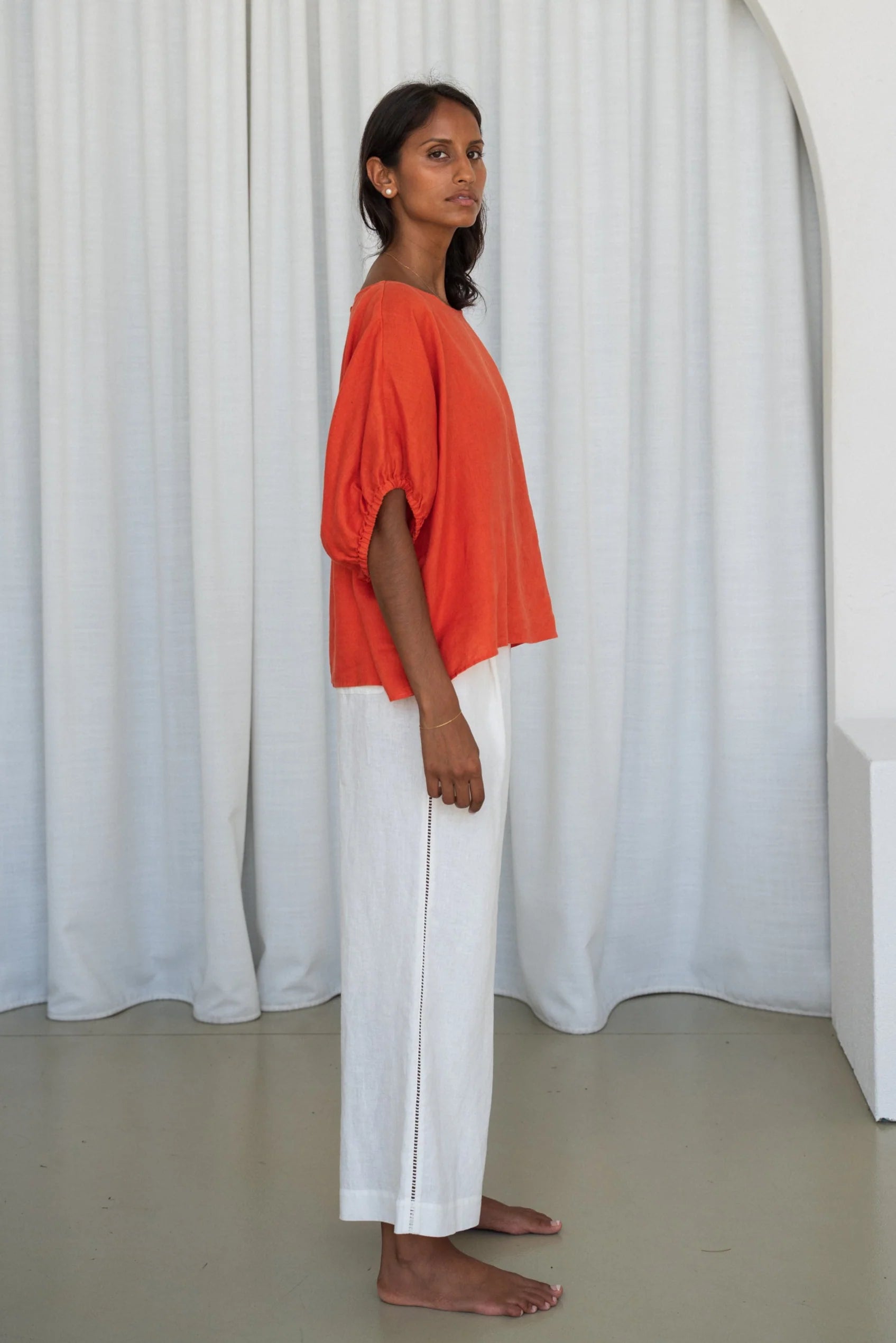 Woman wearing an orange top and white pants standing against a white curtain from Lilly Pilly