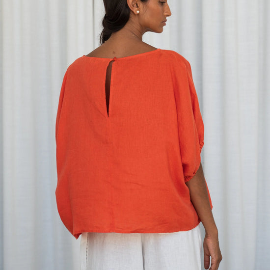 Woman wearing an orange top and white pants against a white curtain background from lilly Pilly