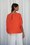 Woman wearing an orange top and white pants against a white curtain background from lilly Pilly