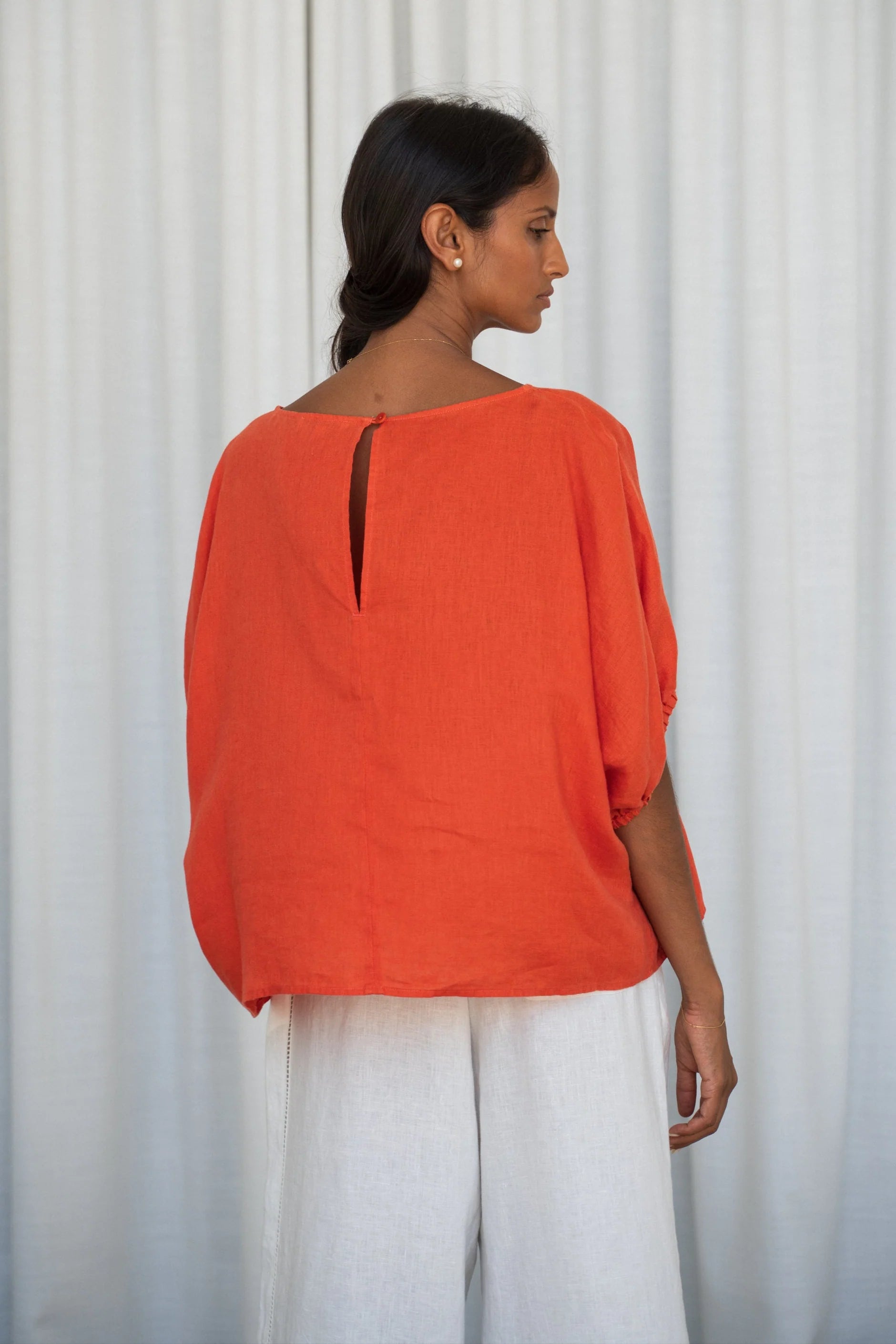 Woman wearing an orange top and white pants against a white curtain background from lilly Pilly