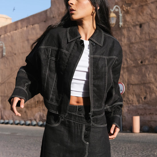 Woman wearing a black linen jacket and black pants standing against a textured wall.