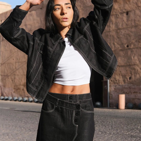 Woman in black linen jacket and skirt posing outdoors against a stone wall.