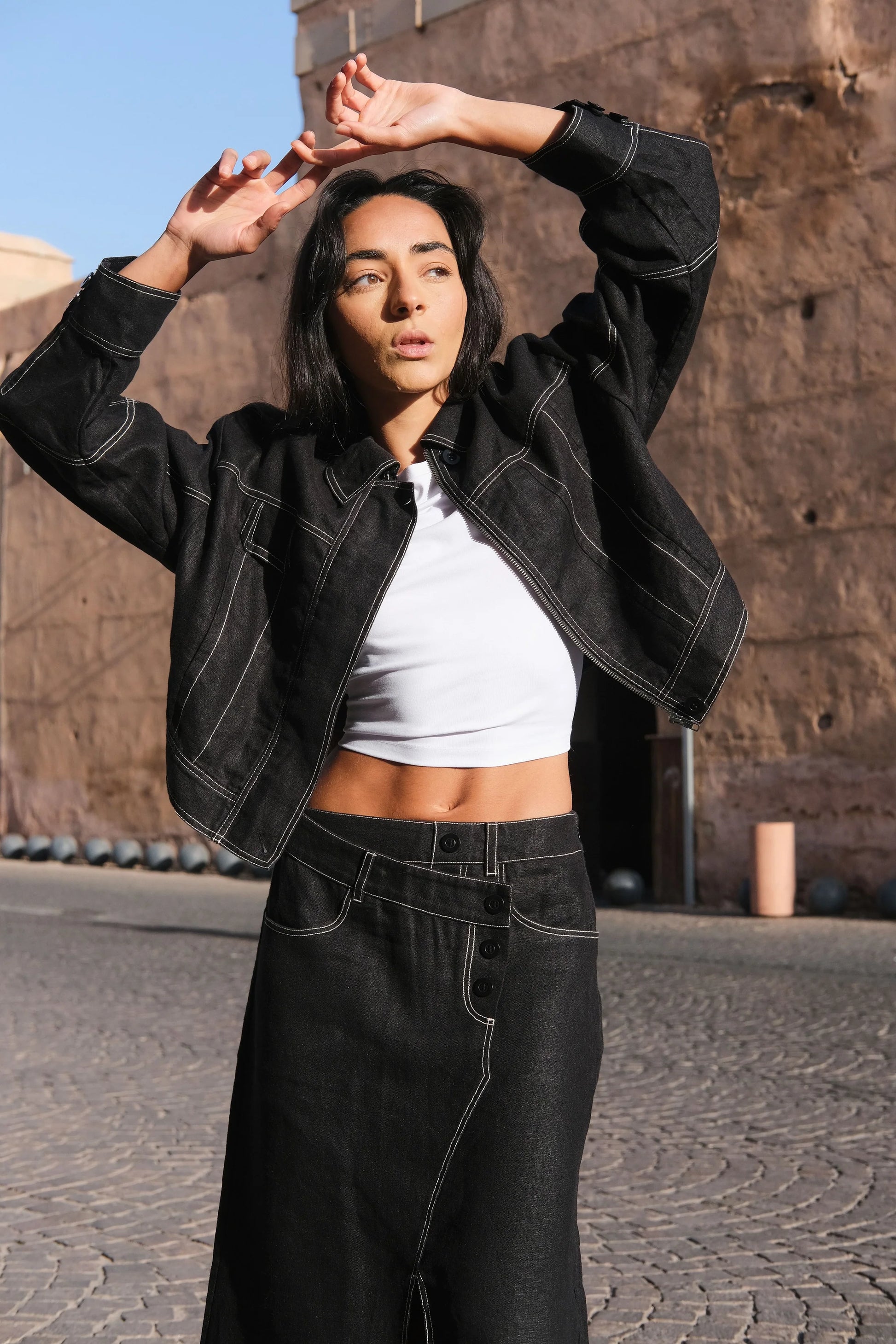 Woman in black linen jacket and skirt posing outdoors against a stone wall.