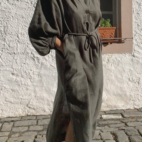 Person wearing a long dark green linen dress standing on a stone pavement.