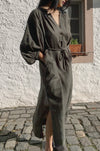 Person wearing a long dark green linen dress standing on a stone pavement.