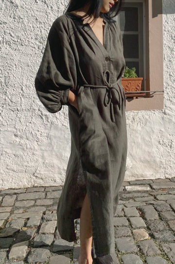 Person wearing a long dark green linen dress standing on a stone pavement.