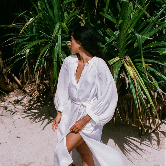 Woman in a white linen dress standing on a sandy beach with green foliage in the background