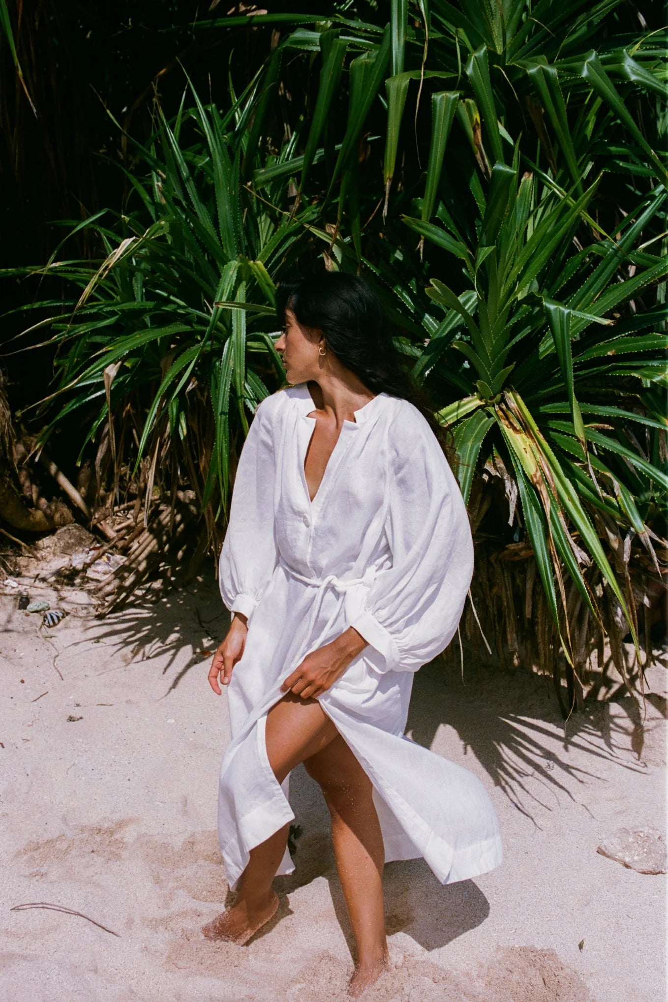 Woman in a white linen dress standing on a sandy beach with green foliage in the background