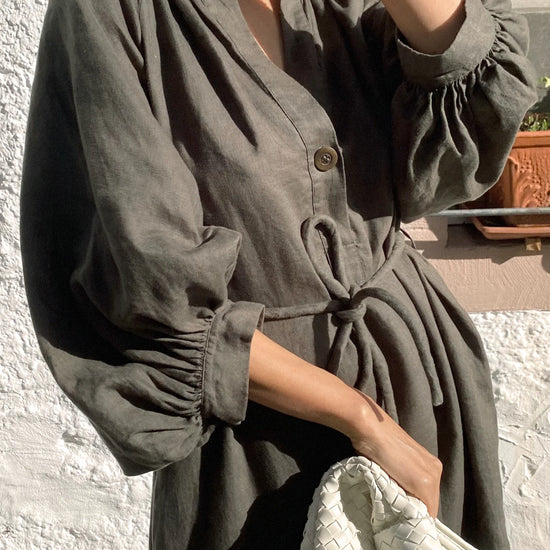 Person wearing a green linen dress holding a white bag against a textured wall.