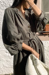 Person wearing a green linen dress holding a white bag against a textured wall.
