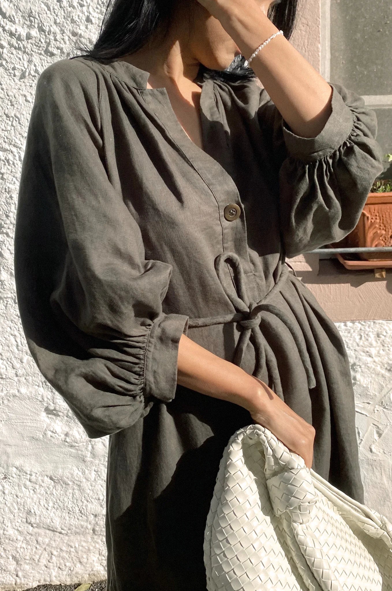 Person wearing a green linen dress holding a white bag against a textured wall.