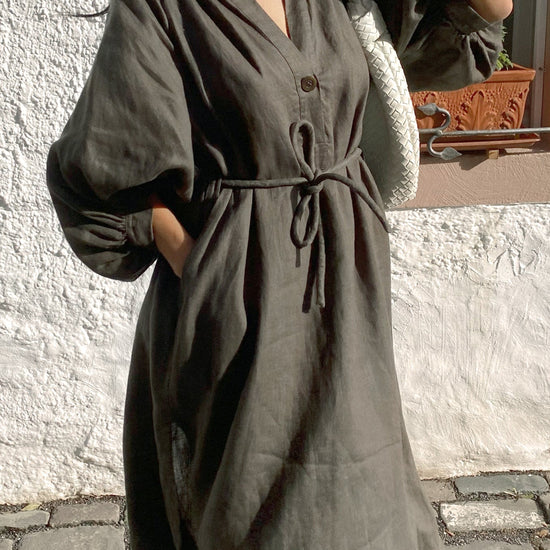 Woman wearing a long, dark green linen dress standing on a stone pavement.
