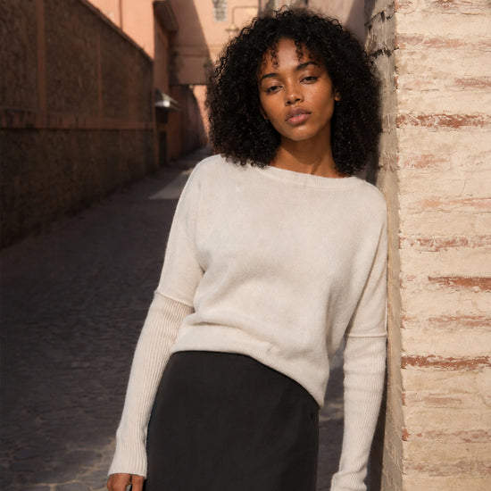 A woman with curly black hair stands in a sunlit outdoor alley wearing a cream-colored knit sweater and a dark skirt.