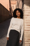 A woman with curly black hair stands in a sunlit outdoor alley wearing a cream-colored knit sweater and a dark skirt.