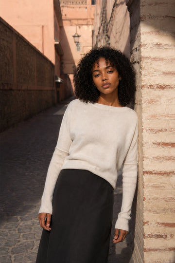 A woman with curly black hair stands in a sunlit outdoor alley wearing a cream-colored knit sweater and a dark skirt.