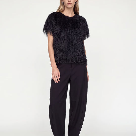 A model stands against a plain light gray studio background wearing a black feather-trimmed short-sleeve top, black wide-leg trousers, and black pointed heels.