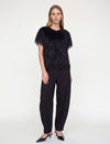 A model stands against a plain light gray studio background wearing a black feather-trimmed short-sleeve top, black wide-leg trousers, and black pointed heels.