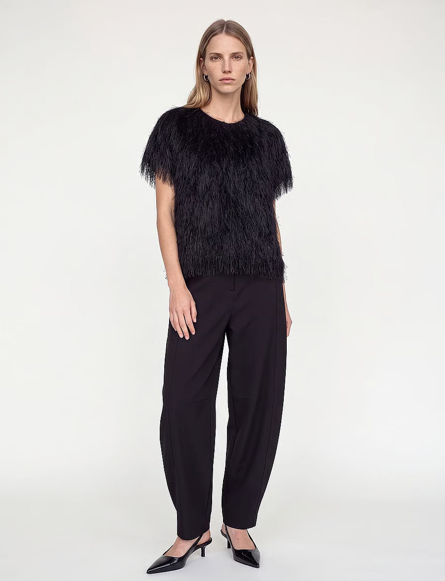A model stands against a plain light gray studio background wearing a black feather-trimmed short-sleeve top, black wide-leg trousers, and black pointed heels.