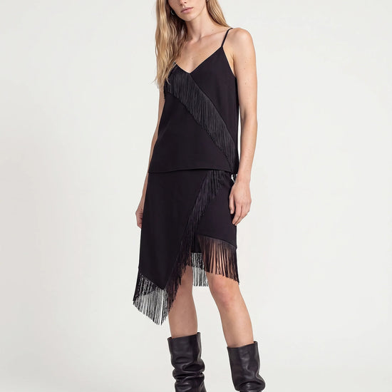 A model wearing a black spaghetti-strap dress with dramatic fringe details and matching fringe boots, photographed against a clean white studio background.