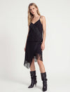 A model wearing a black spaghetti-strap dress with dramatic fringe details and matching fringe boots, photographed against a clean white studio background.