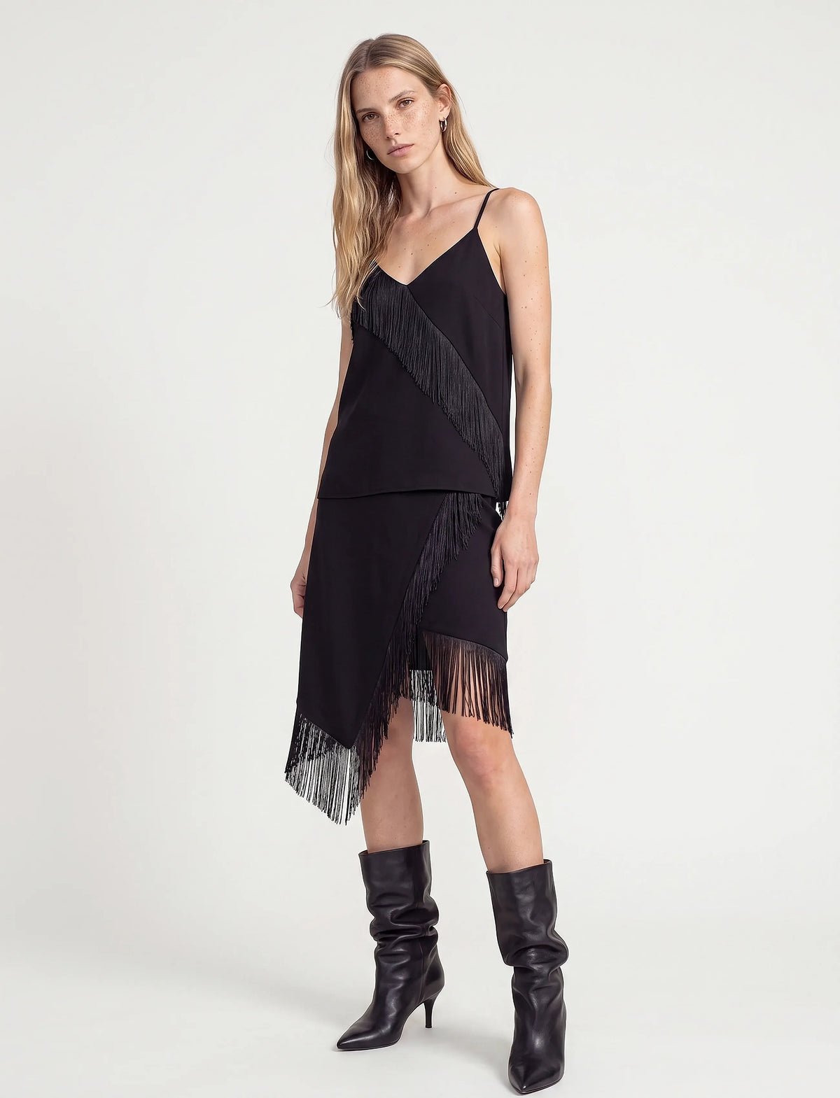 A model wearing a black spaghetti-strap dress with dramatic fringe details and matching fringe boots, photographed against a clean white studio background.