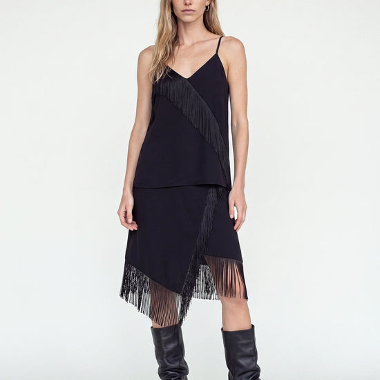 A woman modeling a black sleeveless fringe dress with thin straps and a layered, asymmetrical hem, paired with black knee-high boots against a plain light gray studio background.