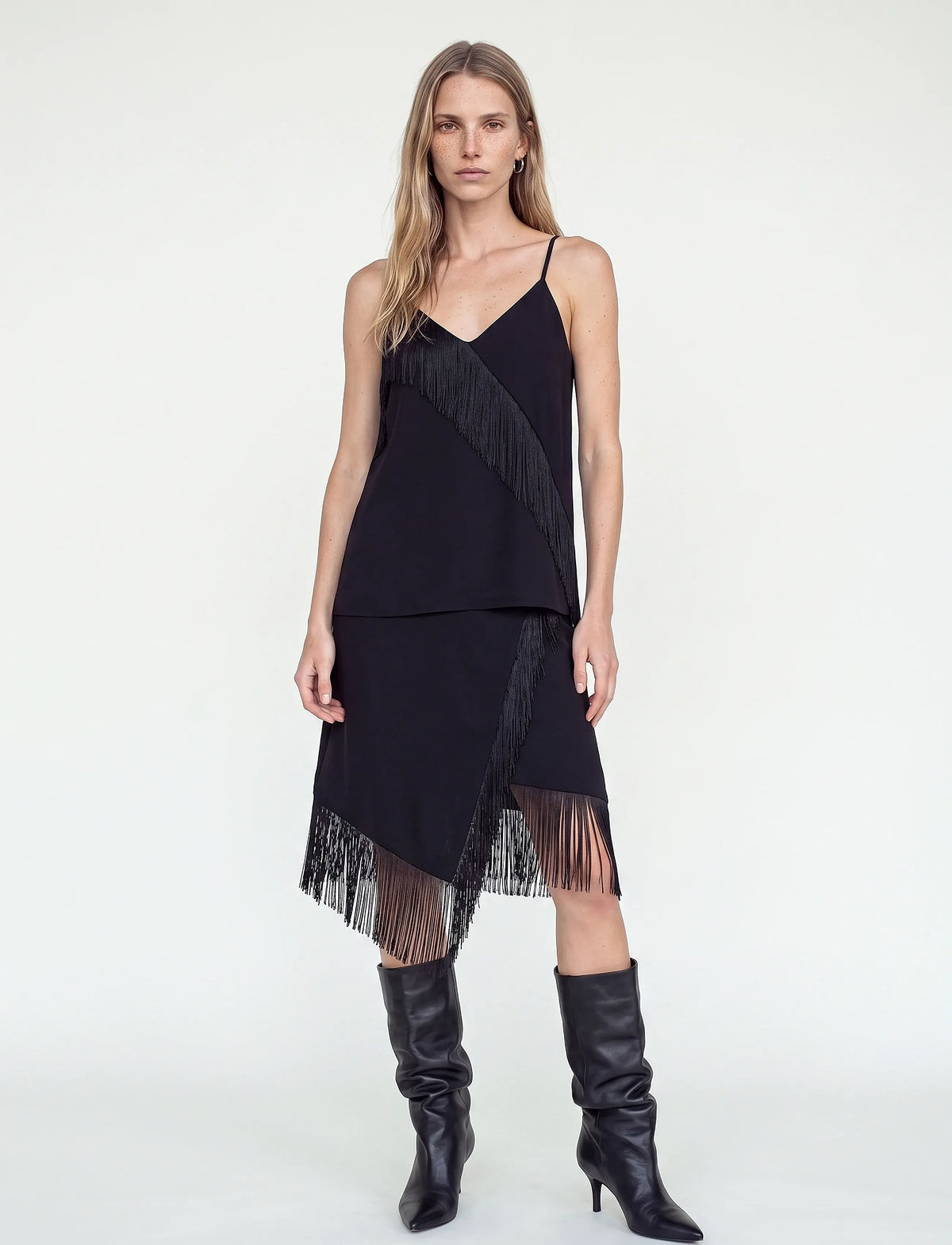 A woman modeling a black sleeveless fringe dress with thin straps and a layered, asymmetrical hem, paired with black knee-high boots against a plain light gray studio background.