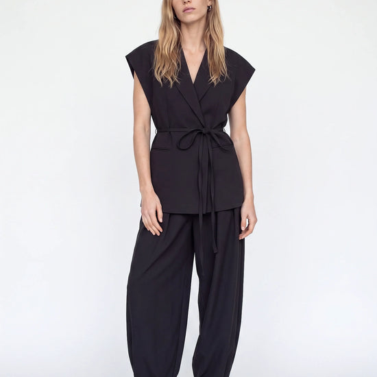 A woman modeling a black two-piece outfit consisting of a sleeveless wrap-style top with a tie waist and matching loose-fitting pants, paired with beige pointed-toe heels against a plain light gray studio background.