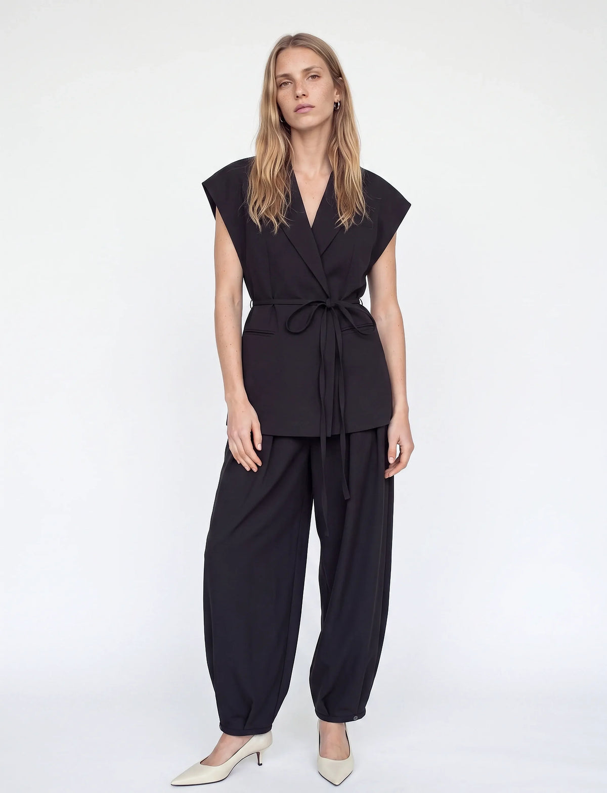 A woman modeling a black two-piece outfit consisting of a sleeveless wrap-style top with a tie waist and matching loose-fitting pants, paired with beige pointed-toe heels against a plain light gray studio background.