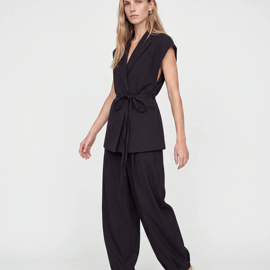 A woman modeling a black two-piece outfit consisting of a sleeveless wrap-style top with a tie waist and matching loose-fitting pants, paired with beige pointed-toe heels against a plain light gray studio background.