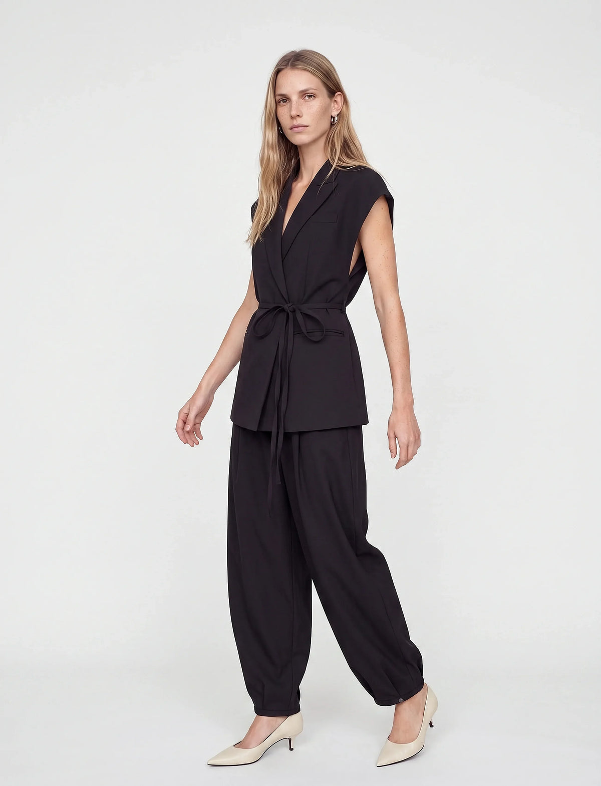 A woman modeling a black two-piece outfit consisting of a sleeveless wrap-style top with a tie waist and matching loose-fitting pants, paired with beige pointed-toe heels against a plain light gray studio background.