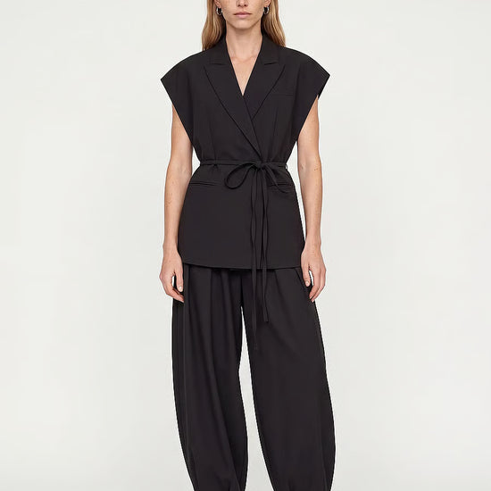 A woman modeling a black sleeveless wrap-style top with a tie waist and matching black wide-leg pants, paired with white pointed-toe heels, against a plain light gray studio background.