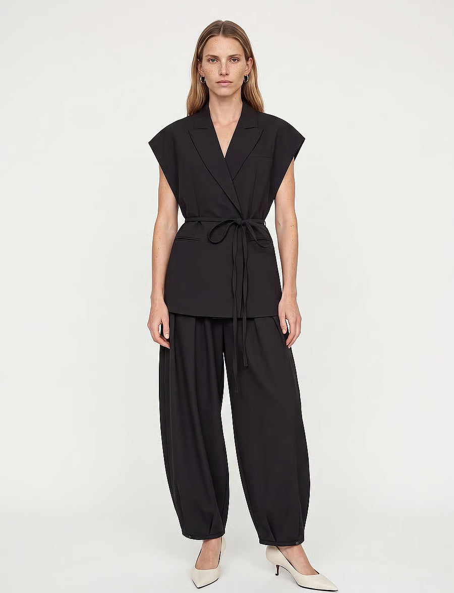 A woman modeling a black sleeveless wrap-style top with a tie waist and matching black wide-leg pants, paired with white pointed-toe heels, against a plain light gray studio background.
