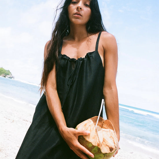 Woman in a black dress holding a coconut on a beach