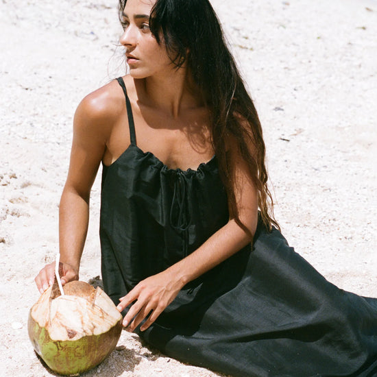 Woman in a black dress sitting on a sandy beach with a coconut.