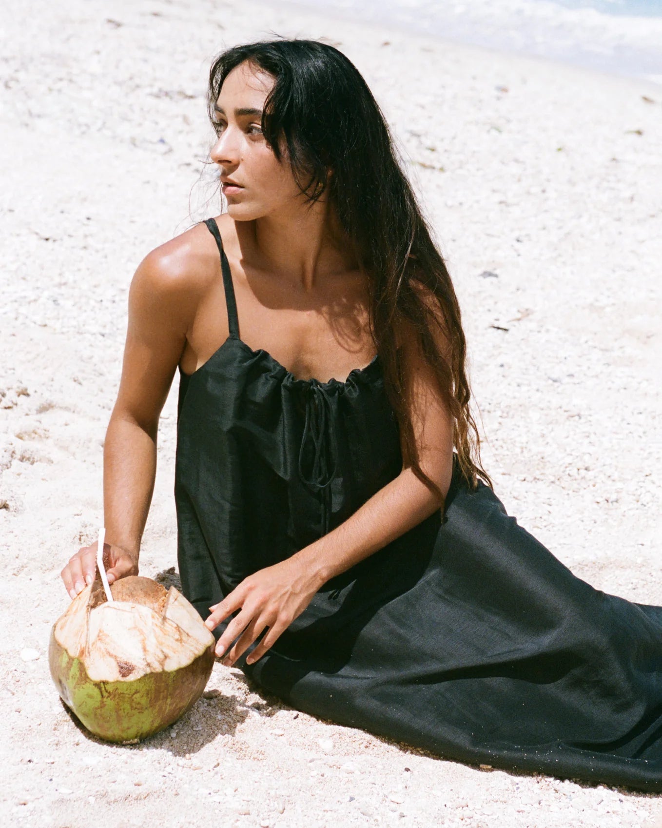 Woman in a black dress sitting on a sandy beach with a coconut.