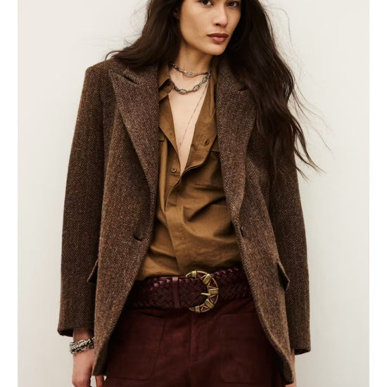 A model wearing a brown textured tweed blazer over a brown button-up shirt, paired with dark burgundy trousers and a dark brown braided belt with a gold buckle, set against a plain off-white background.