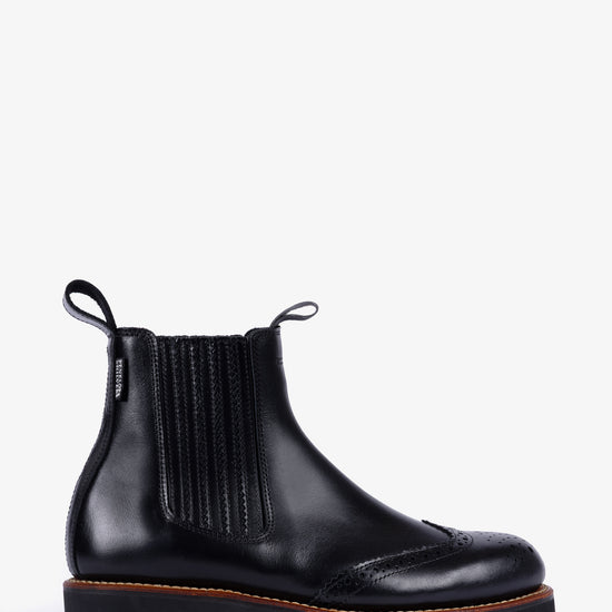 A black leather Chelsea boot with brogue detailing on the toe, elastic side panels, and a chunky lug sole, shown from a side profile against a light gray background.