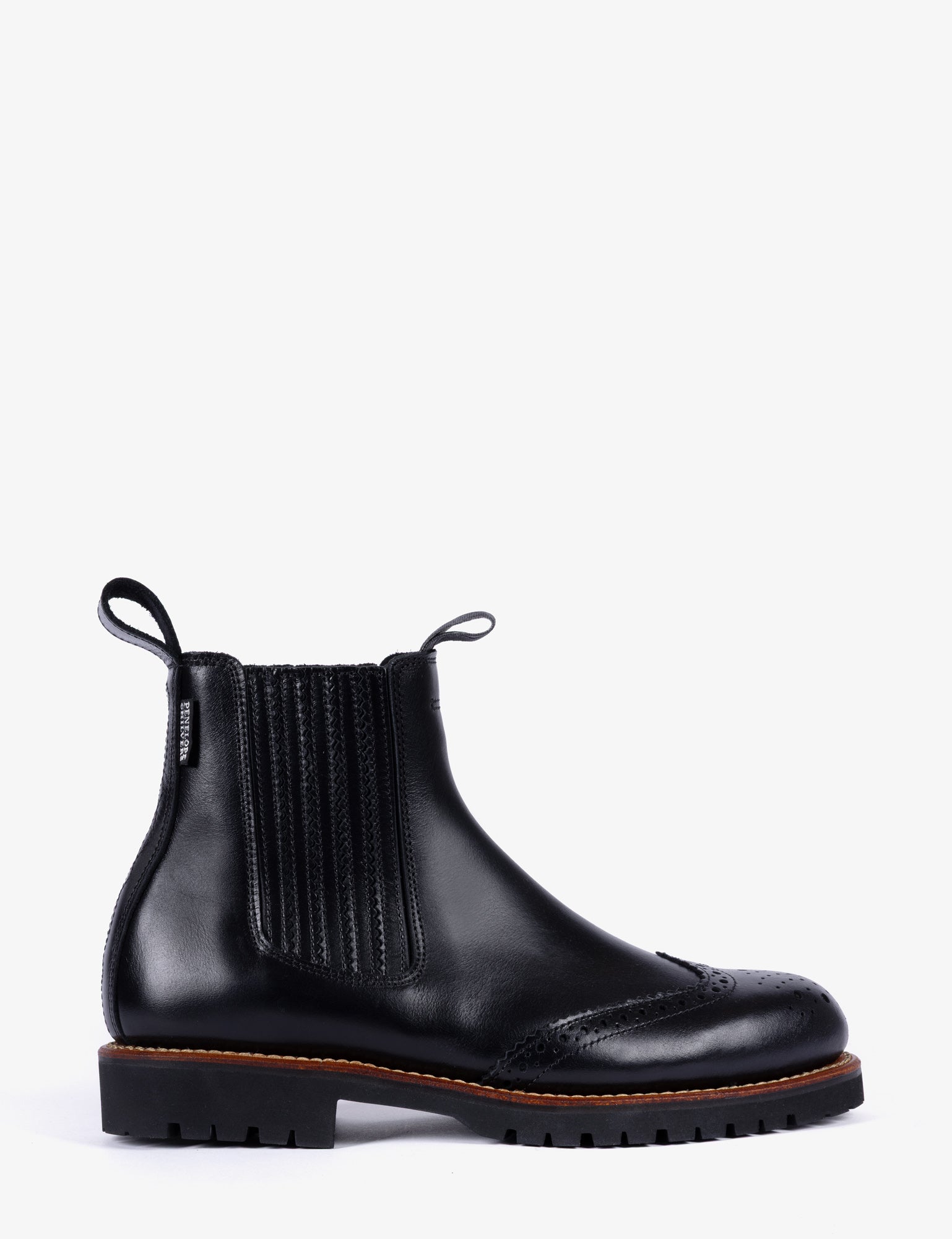 A black leather Chelsea boot with brogue detailing on the toe, elastic side panels, and a chunky lug sole, shown from a side profile against a light gray background.