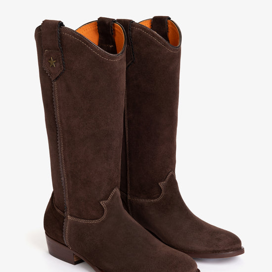 A pair of dark brown suede cowboy boots with a low block heel and a small gold star detail on the pull tab, shown against a plain white background.