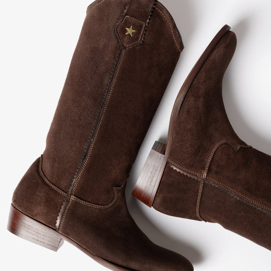 A pair of dark brown suede cowboy boots with a small gold star detail on the shaft, shown from a high-angle studio perspective on a white background.