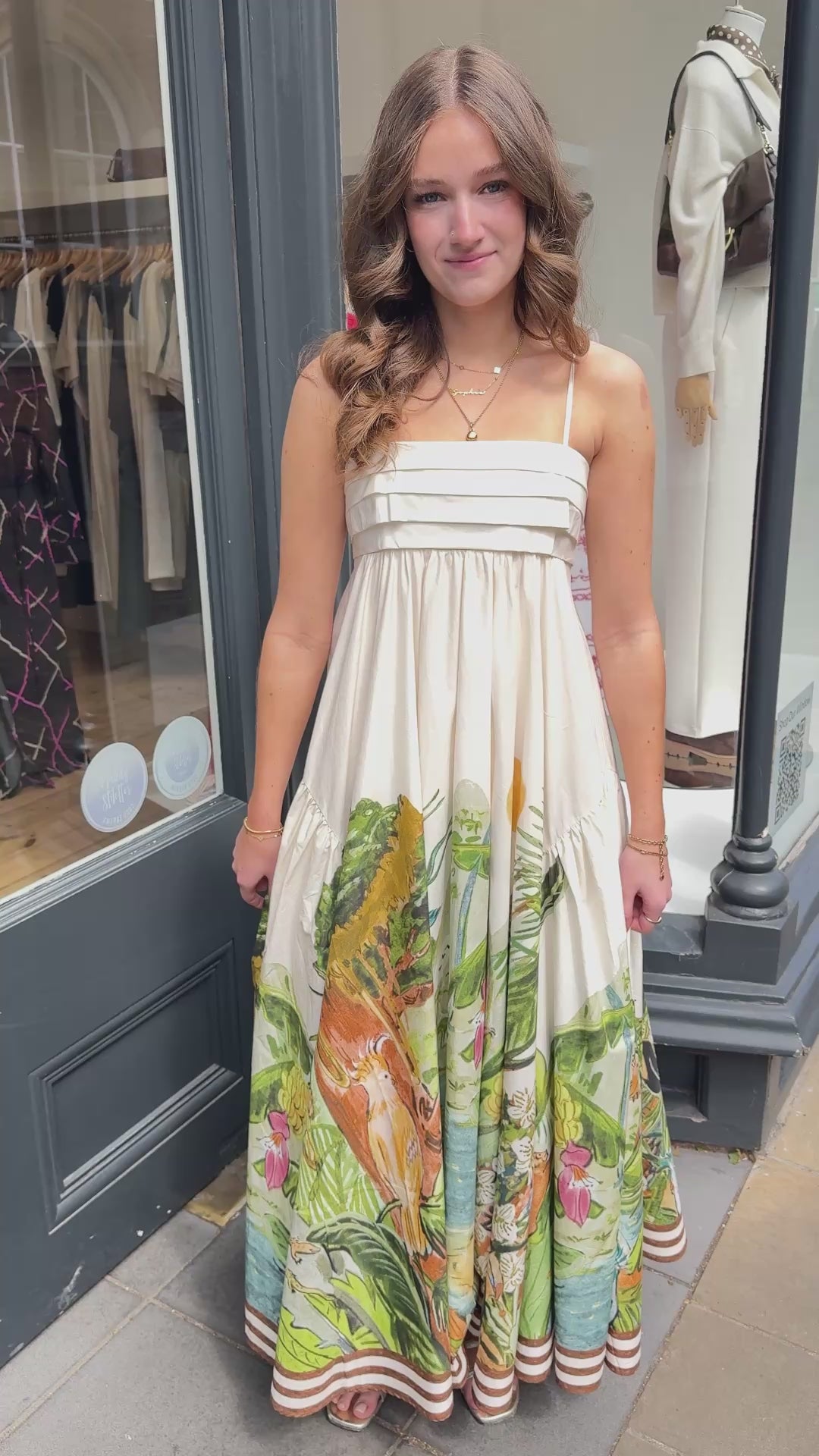 Model wearing cream sundress with jungle print at the bottom standing in front of a shop video. 