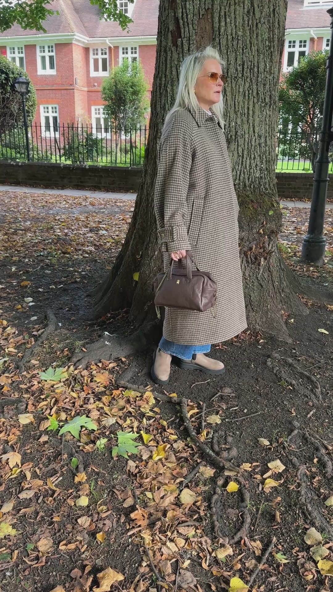Model video in autumnal scenery wearing  a long wool houndstooth coat holding brown leather handbag with gold scarab stamp.